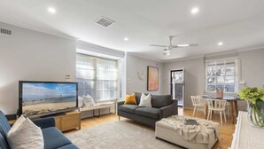 A modern living room with a flat-screen TV, a grey sofa, a blue sofa, a grey armchair, a small table with chairs, and a ceiling fan.