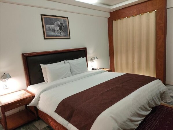 Une chambre d’hôtel avec un grand lit, une table de chevet avec une lampe, un tableau encadré au mur et une fenêtre avec des rideaux.