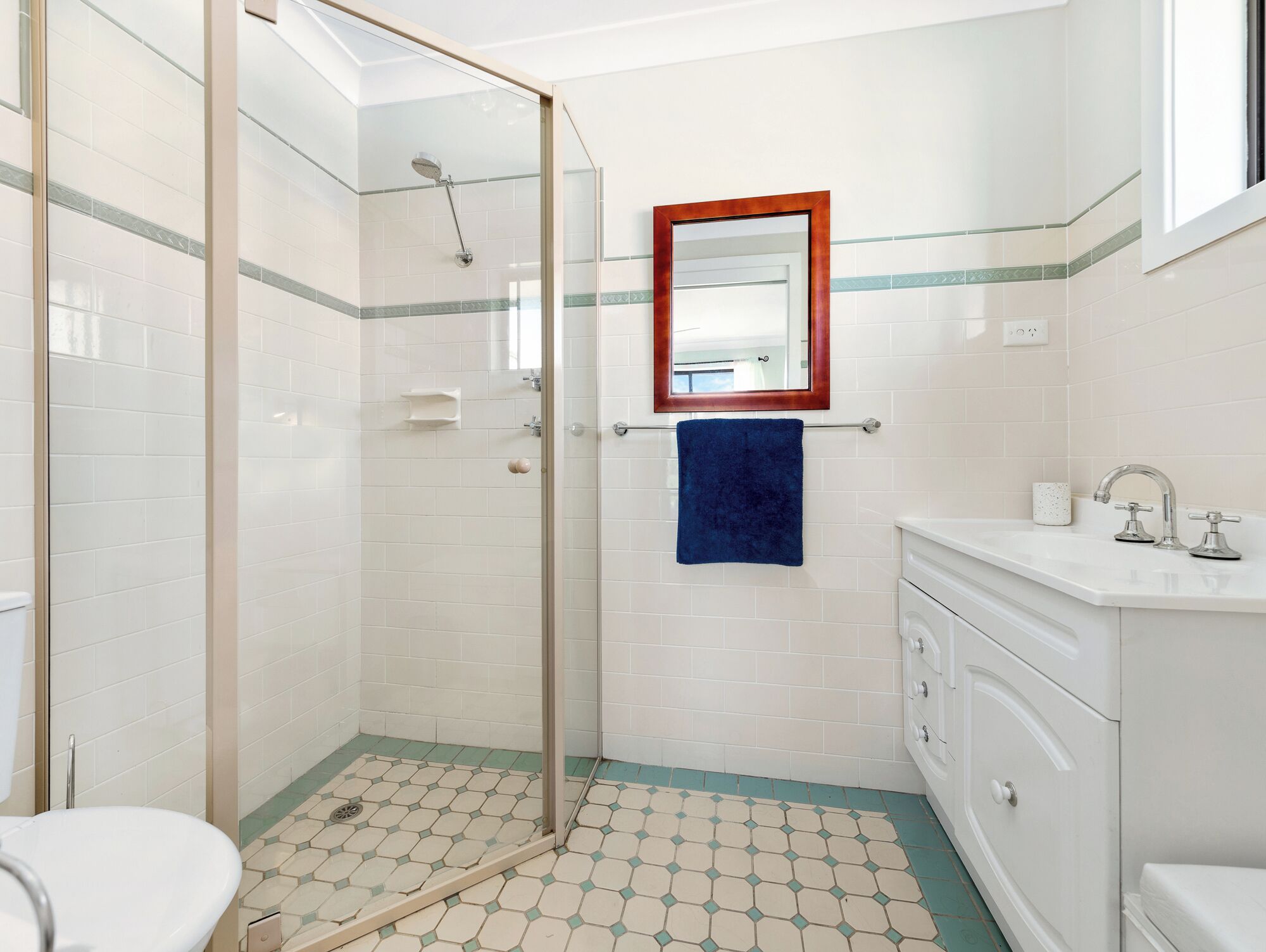 A bathroom with a glass shower enclosure, a white sink with a mirror above it, and a blue towel hanging on the wall.