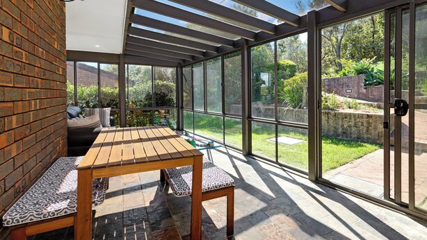 Une terrasse ensoleillée avec une table et des bancs en bois, de grandes portes vitrées et un mur en briques.