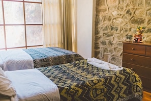 A room with two beds, a stone wall, and a dresser with a teddy bear. - Hostal Aylluq Wasin  (Urubamba)