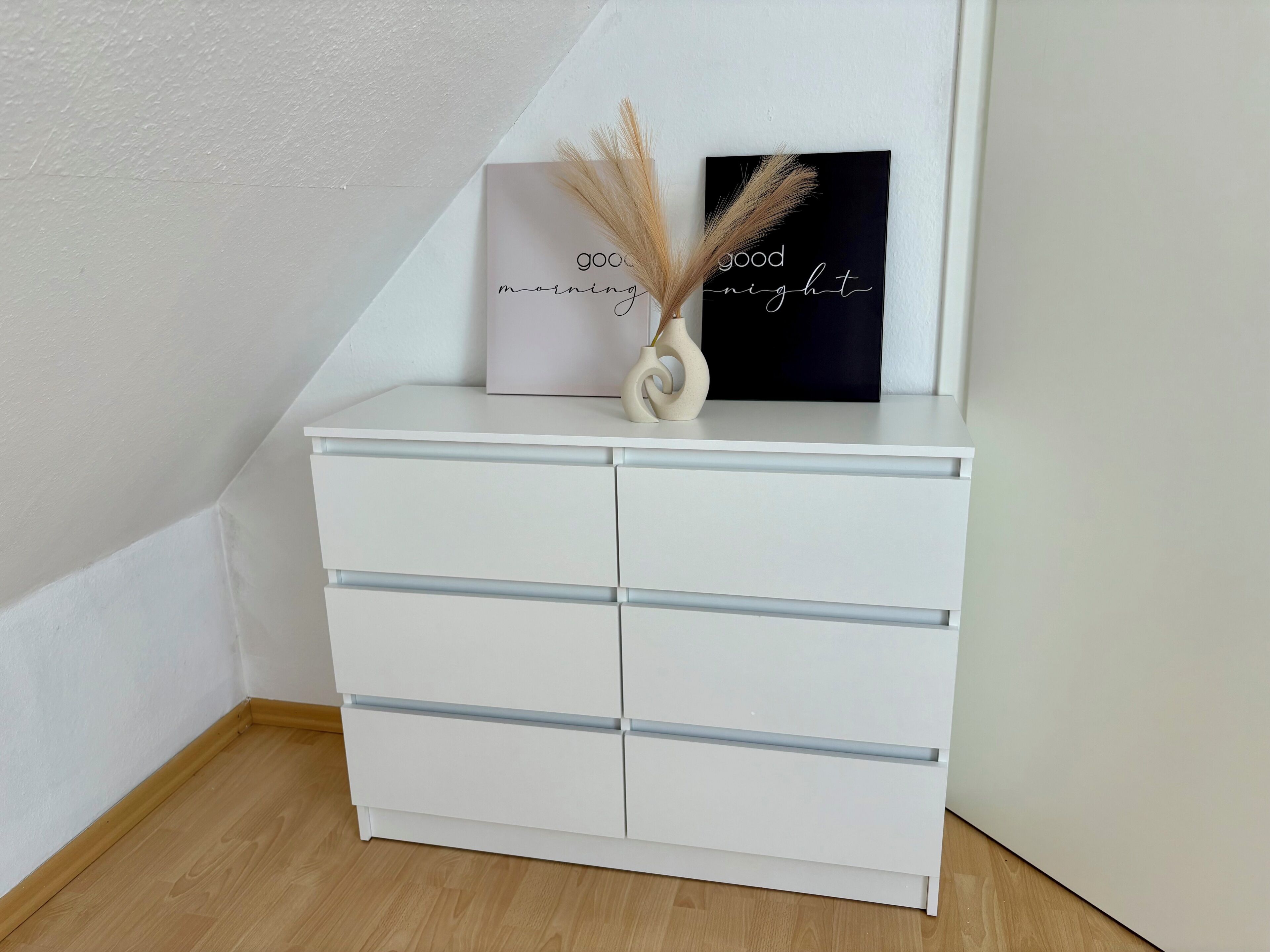A white chest of drawers with a vase of dried pampas grass and a framed sign with the text 'good morning night' on a wall.