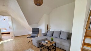 Une chambre de style loft moderne, avec un plafond en pente, un canapé gris, un fauteuil à bascule en bois, une petite table basse en bois et une porte blanche donnant sur une autre pièce.