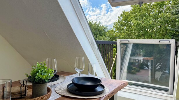 Un coin repas avec une table dressée pour deux personnes, une plante en pot et une vue sur le ciel et les arbres, visible par une fenêtre.