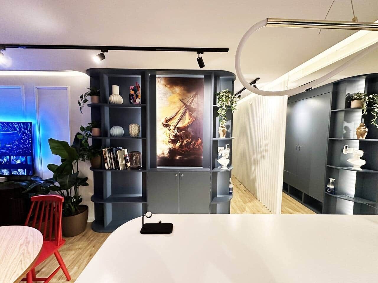 Een moderne woonkamer met een grote flatscreen televisie, een eetgedeelte met een witte tafel en rode stoelen, en een grijze boekenkast met decoratieve objecten.