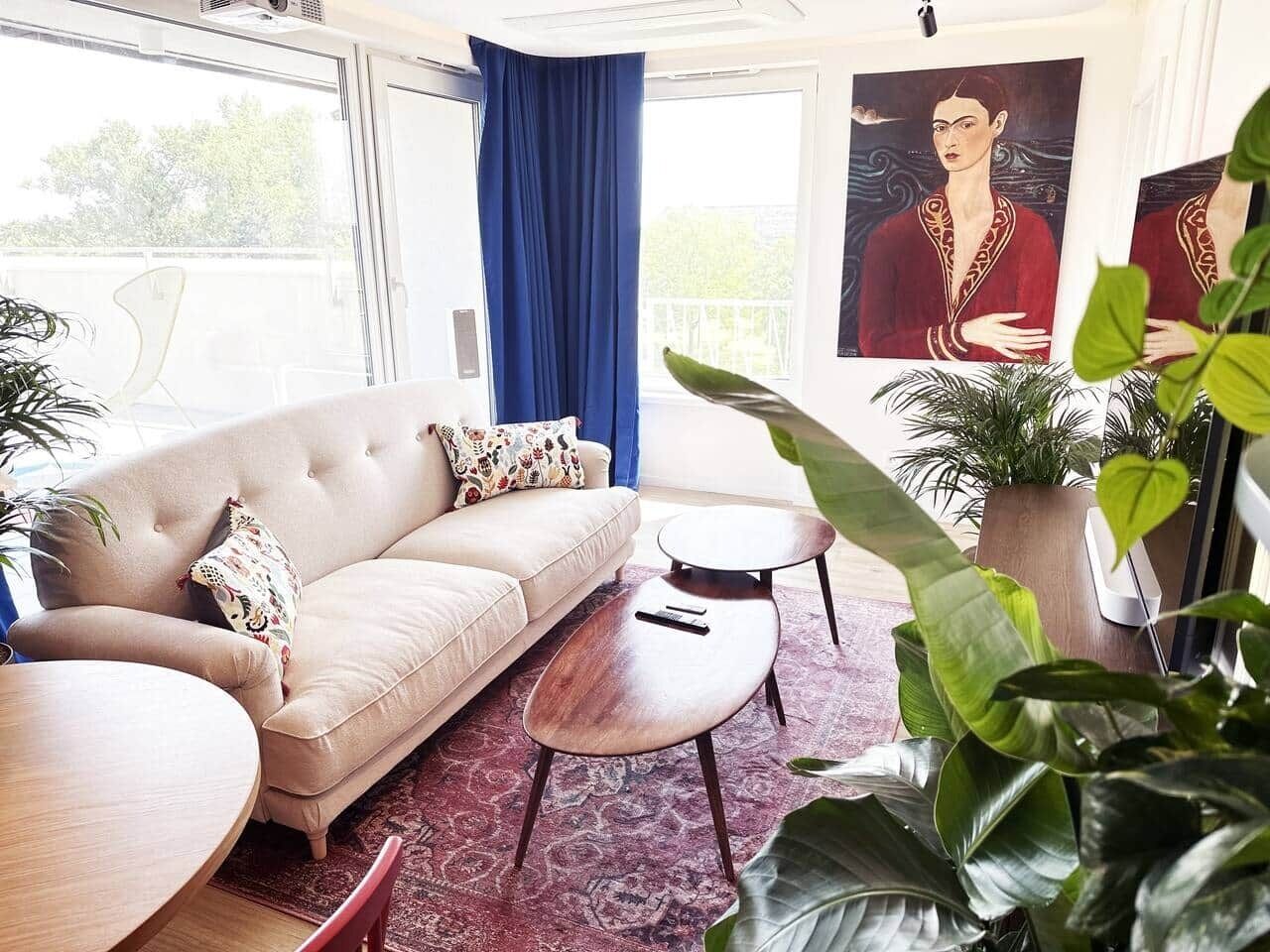 A living room with a sofa, coffee table, and chairs, a large window with curtains, and indoor plants.