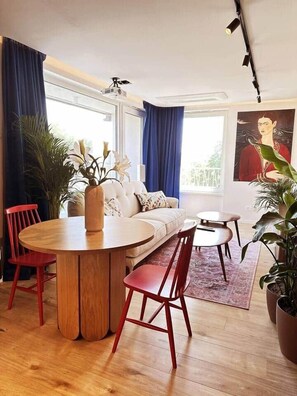 A modern living room with a round wooden dining table, red chairs, a sofa, and a large window with blue curtains.