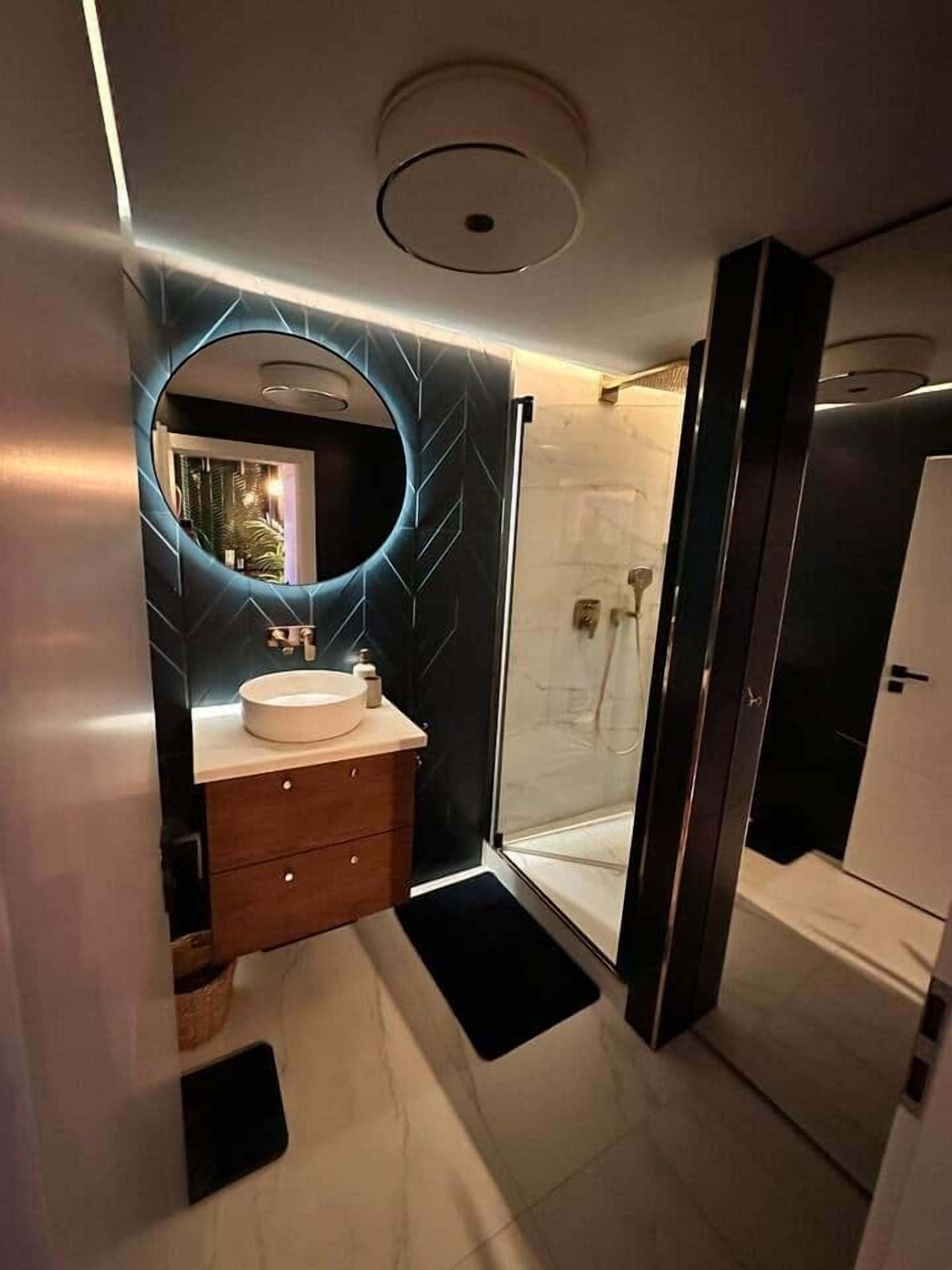 Ein modernes Badezimmer mit rundem Spiegel, Waschbecken in Weiß und einer Duschkabine aus Glas.