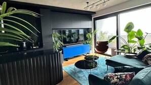 Un salon moderne avec un canapé bleu, une table basse noire et un téléviseur posé sur un meuble bleu. On y trouve plusieurs plantes en pot et une grande fenêtre qui laisse entrer la lumière naturelle.