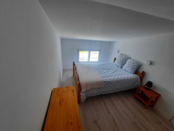 A small, single-bed room with a sloped ceiling, a wooden bench, a small table, and a window providing natural light.