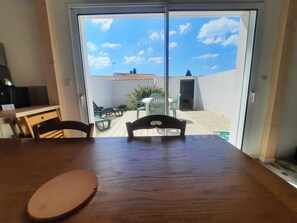 Quarto com uma mesa e cadeiras de madeira, porta de vidro deslizante que dá para um pátio externo com mesa e cadeiras, e uma pequena cozinha com geladeira e pia.