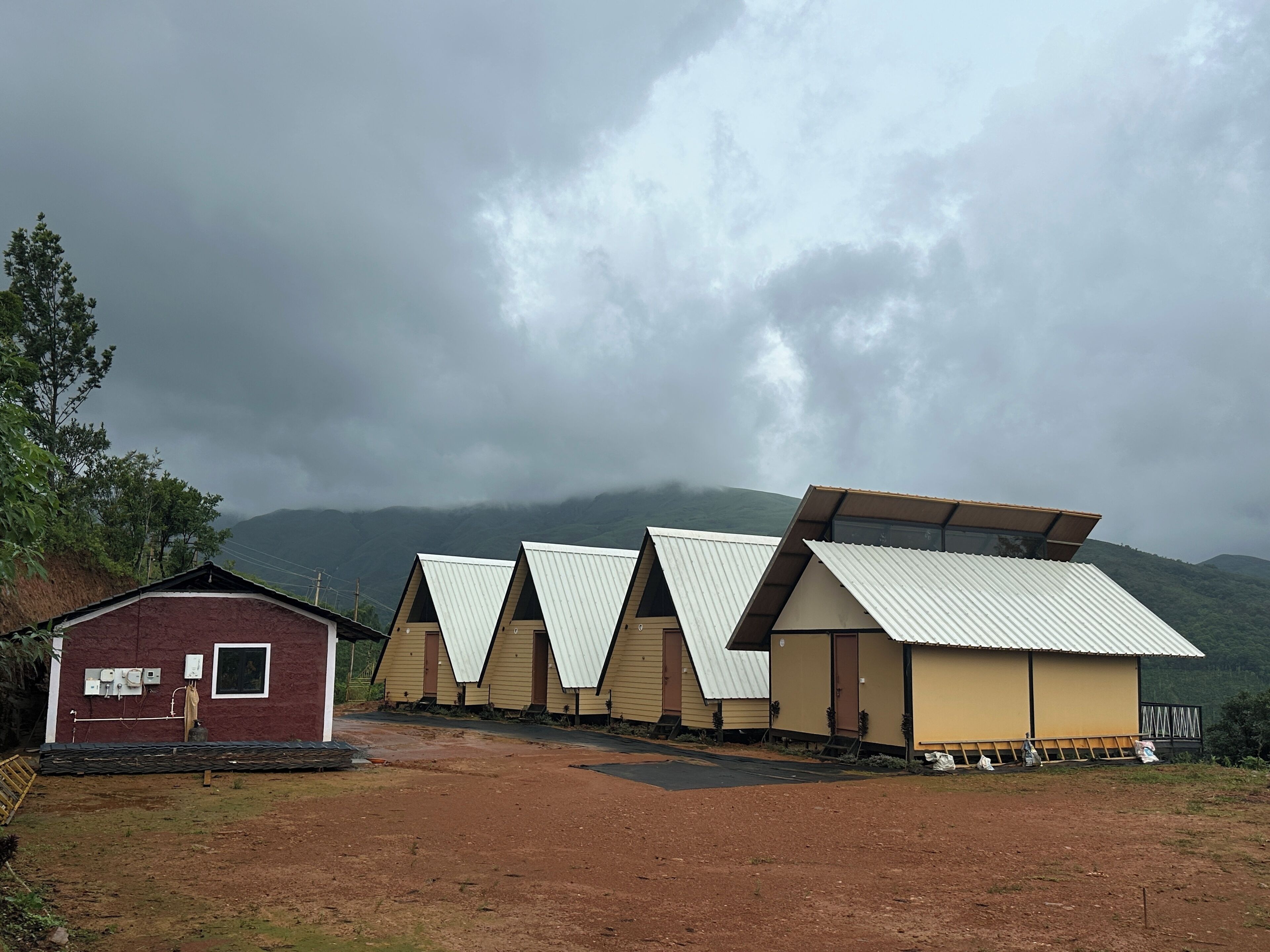 A row of triangular-roofed cabins with a dirt path leading to them, set against a backdrop of mountains and cloudy skies.
