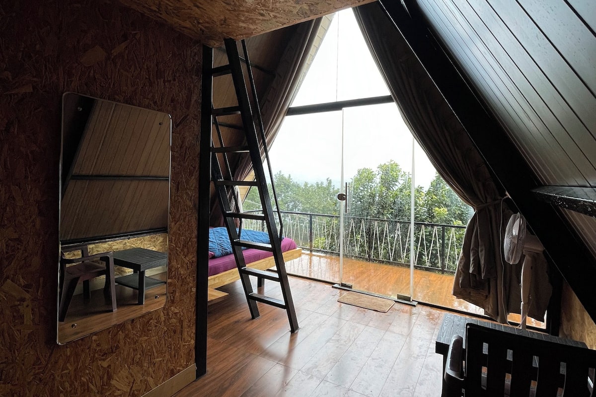 A loft bedroom with a sloped ceiling, wooden flooring, a ladder, a bed, a small table, and a large window offering a view of greenery.