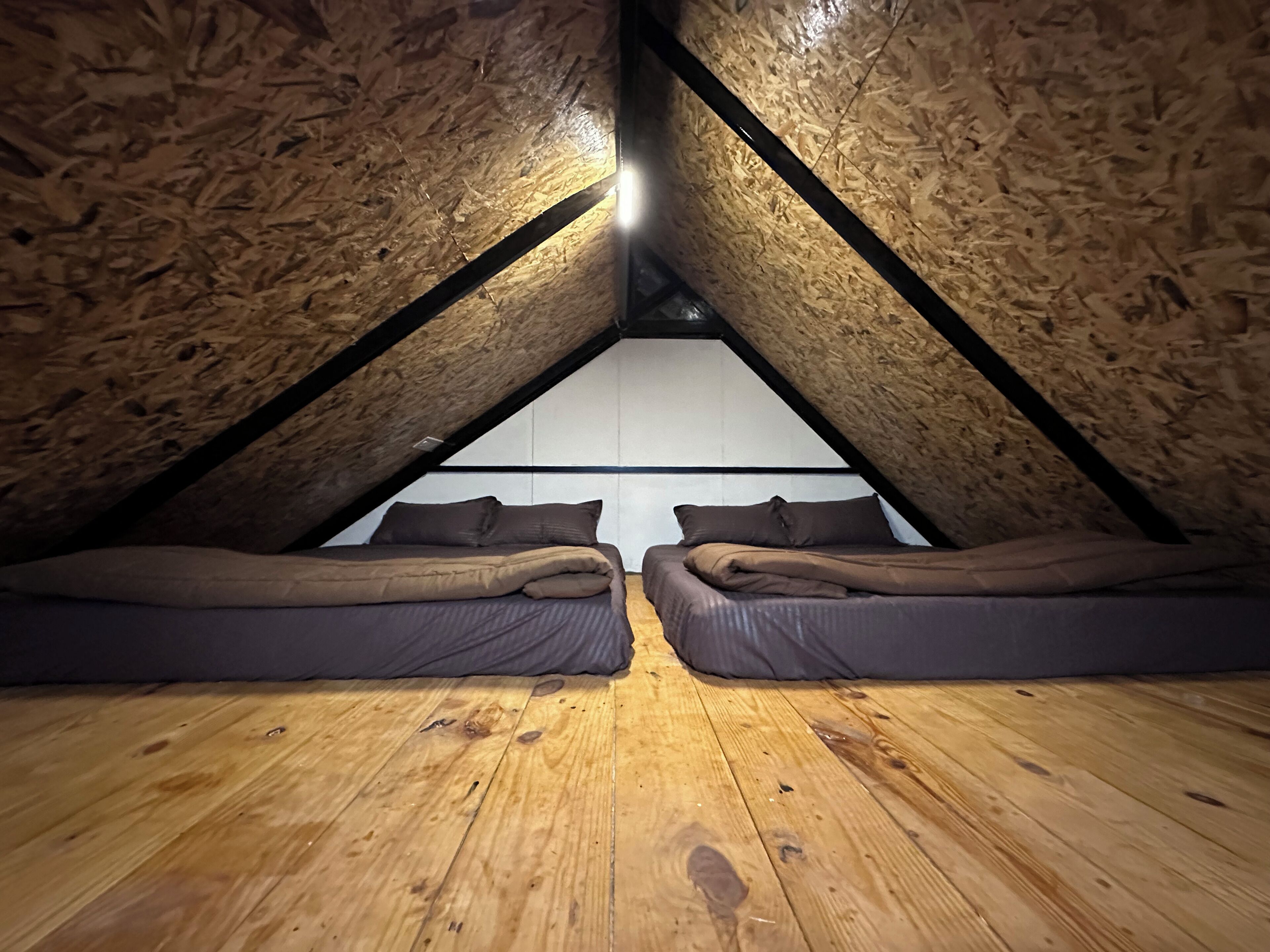 A small attic bedroom with wooden walls and a sloped ceiling. There are two beds with dark bedding and a wooden floor.