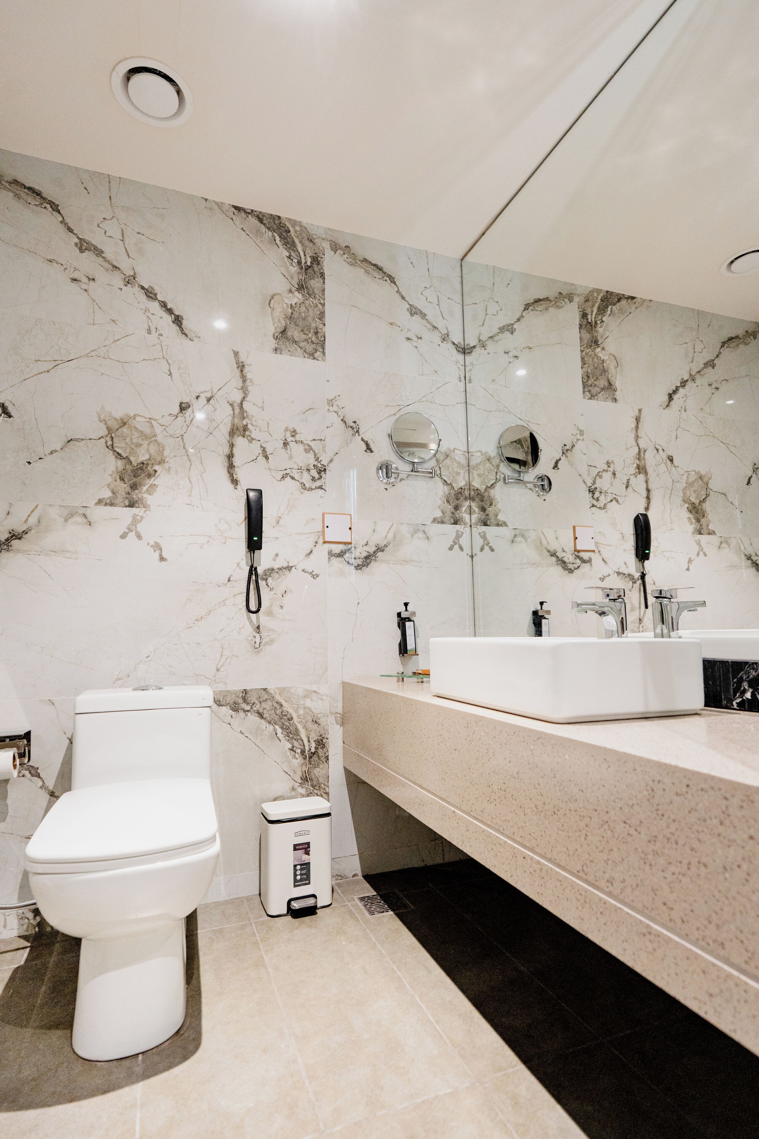 A modern bathroom with a marble wall, a white toilet, a sink, and a trash bin.