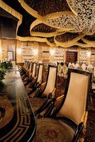 Uma sala de banquetes com uma mesa comprida, cadeiras ornamentadas e detalhes decorativos no teto.