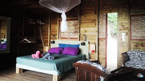 A wooden cabin bedroom with a bed, a chair, a hanging mosquito net, and a window.