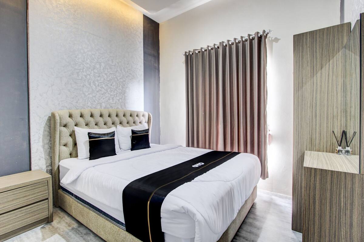 Una habitación de hotel moderna con una cama de cabecera acolchada, una mesita de noche de madera, un espejo grande y una ventana con persianas verticales.