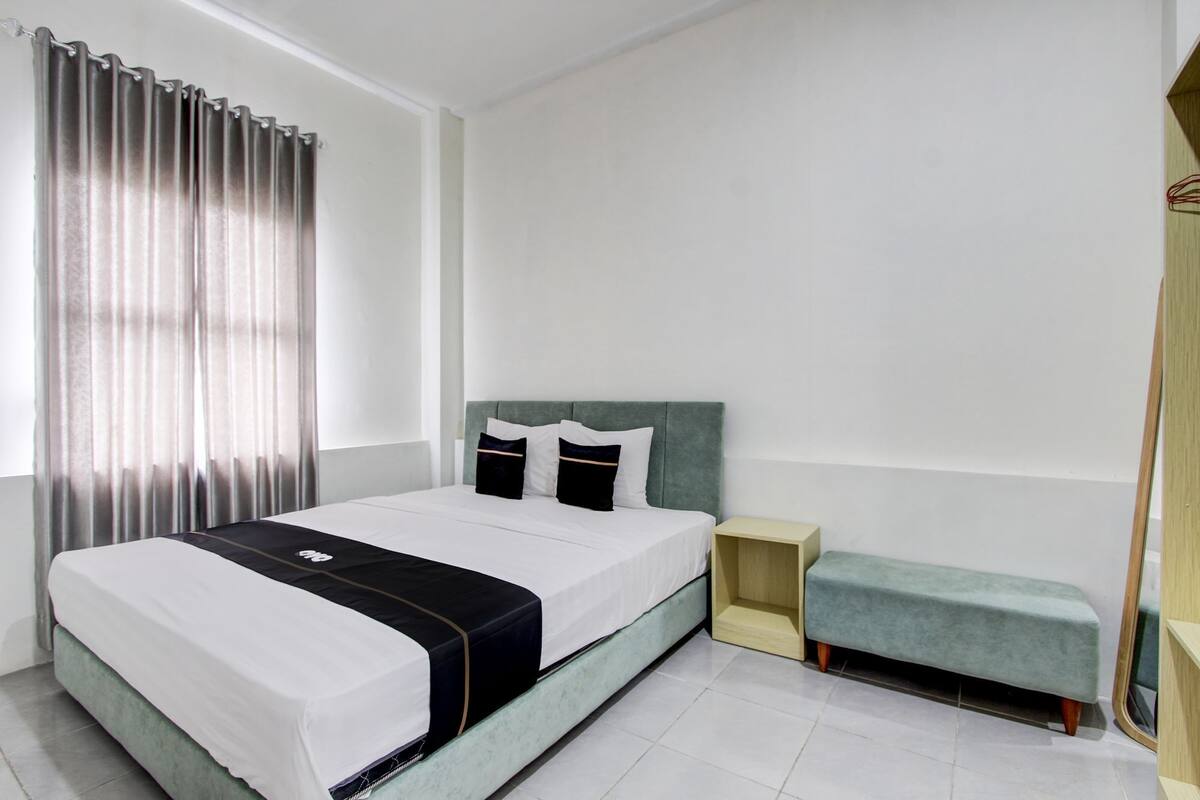 Un dormitorio moderno con cama, mesita de noche, banco y un amplio ventanal con cortinas.
