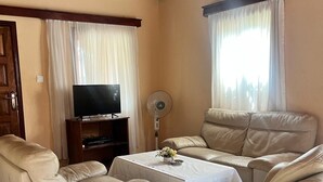 A living room with a sofa set, a television, a fan, and a table with flowers. - Ocean Walk Guesthouse (Brufut)