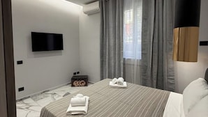 A modern hotel room with a flat-screen TV mounted on the wall, a bed with striped bedding, a small wooden chest, and a window with grey curtains. - Feffo's home (Roma)