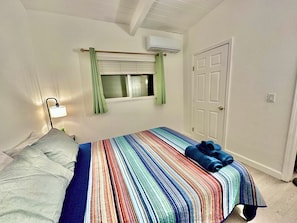 A bedroom with a bed, a nightstand, a lamp, a window with curtains, an air conditioning unit, and a door.