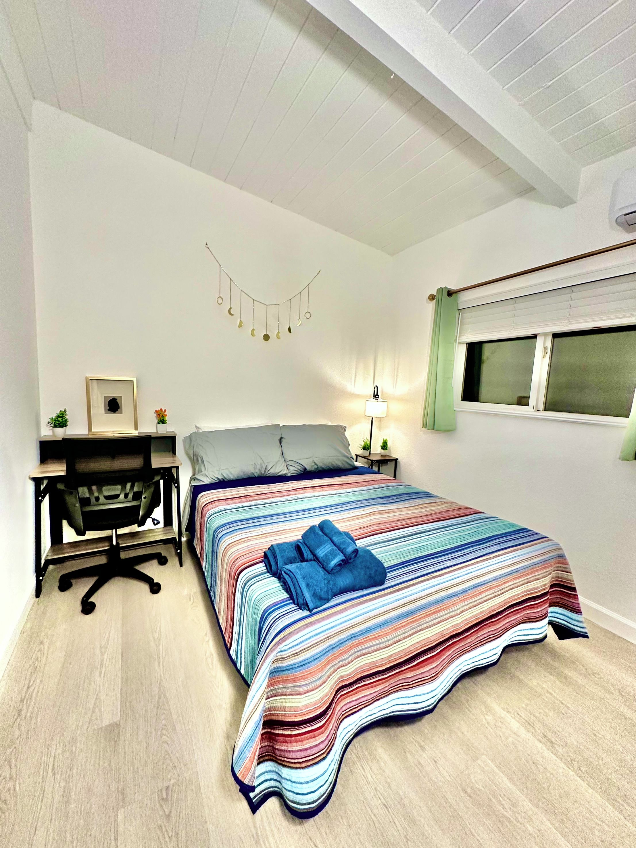 A bedroom with a bed, a desk, a chair, a lamp, and a window with blinds.