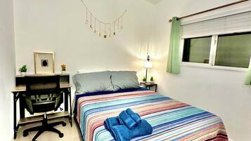 A bedroom with a bed, a desk, a chair, a lamp, and a window with blinds.