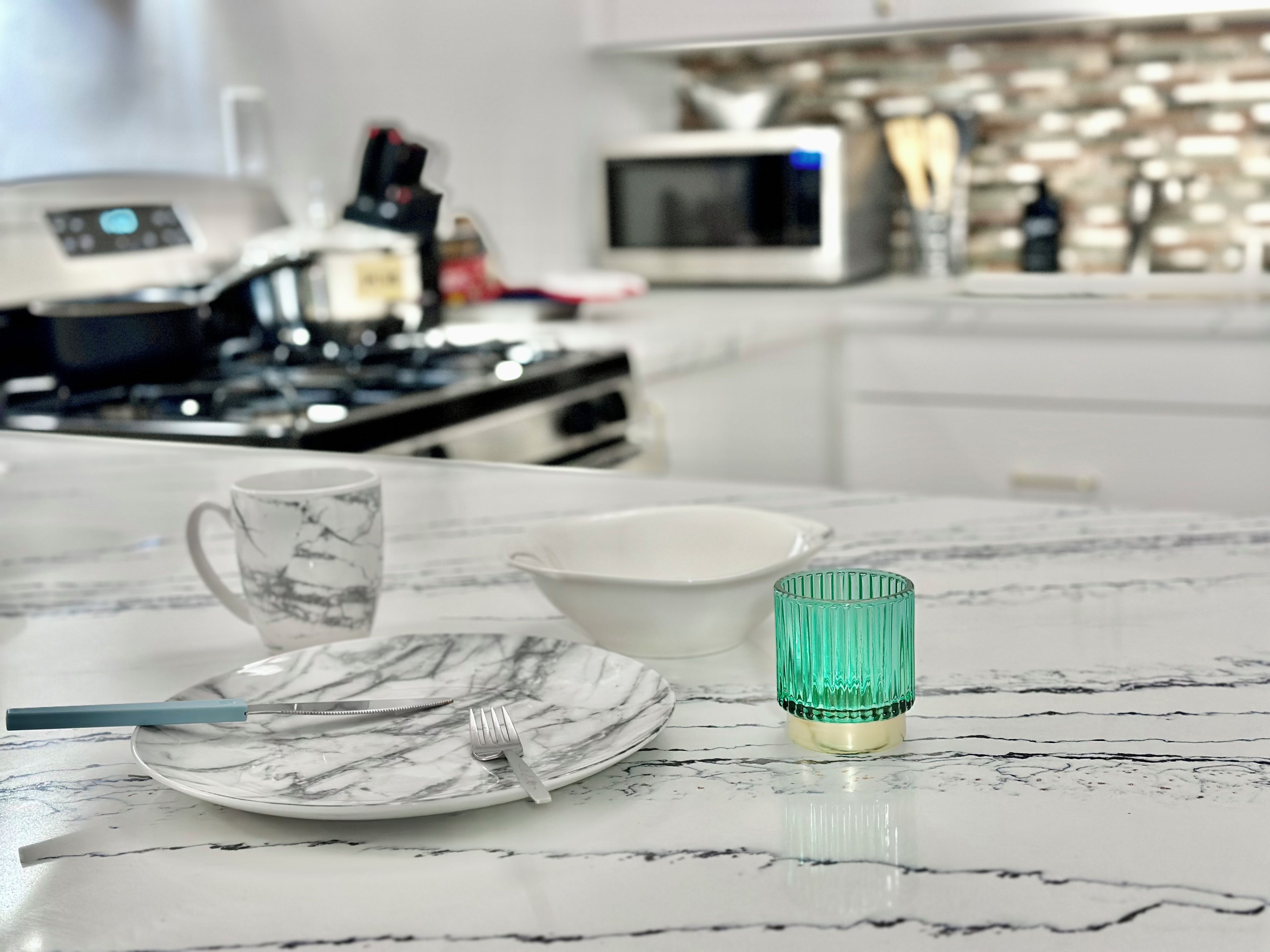 A kitchen counter with a white marble surface, a white plate, a white bowl, a green glass, a blue knife, and a silver fork.