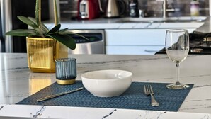 A modern kitchen with a marble countertop, a white bowl, a glass of water, a fork, a knife, a blue mat, a gold vase, and a green plant.
