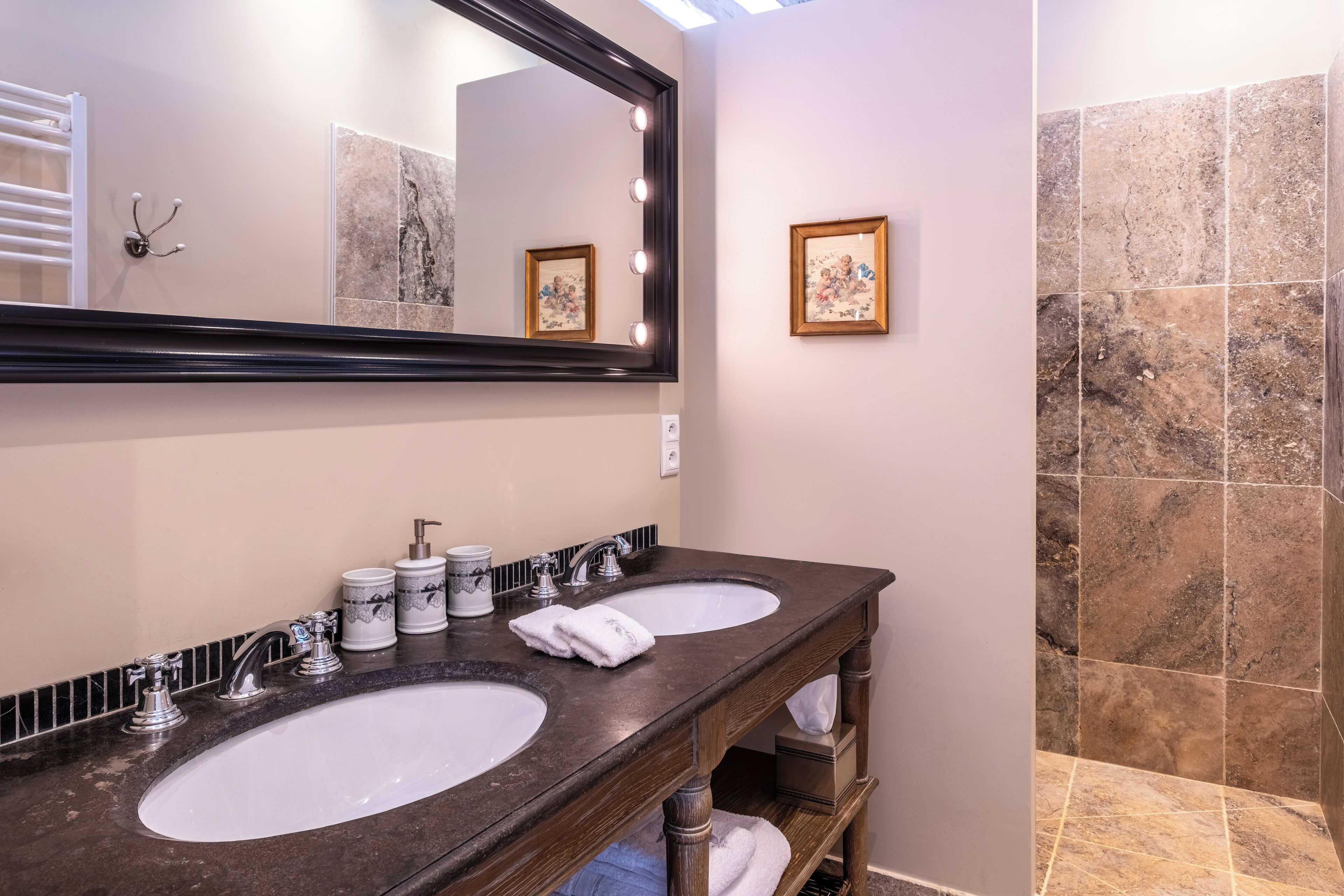 A bathroom with two sinks, a large mirror with lights, a towel rack, and a framed picture on the wall.