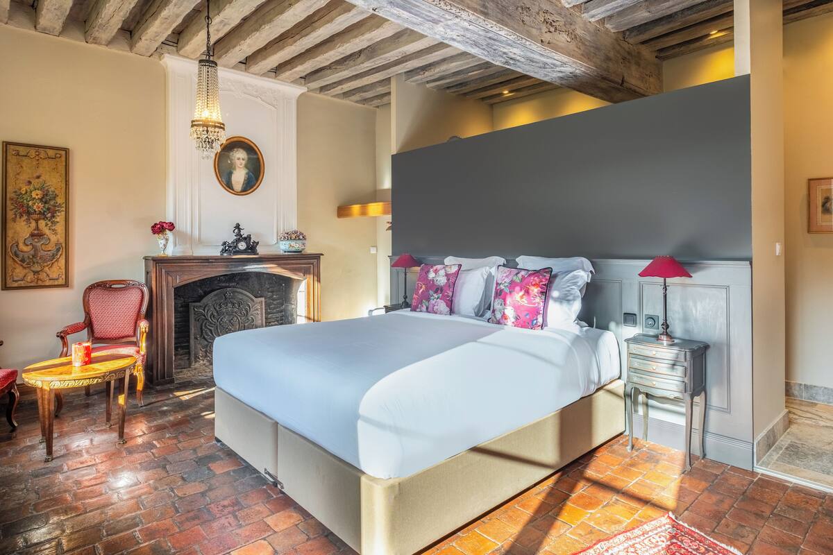 Een ruime slaapkamer met een groot bed, een open haard, een kroonluchter en een rode stoel.