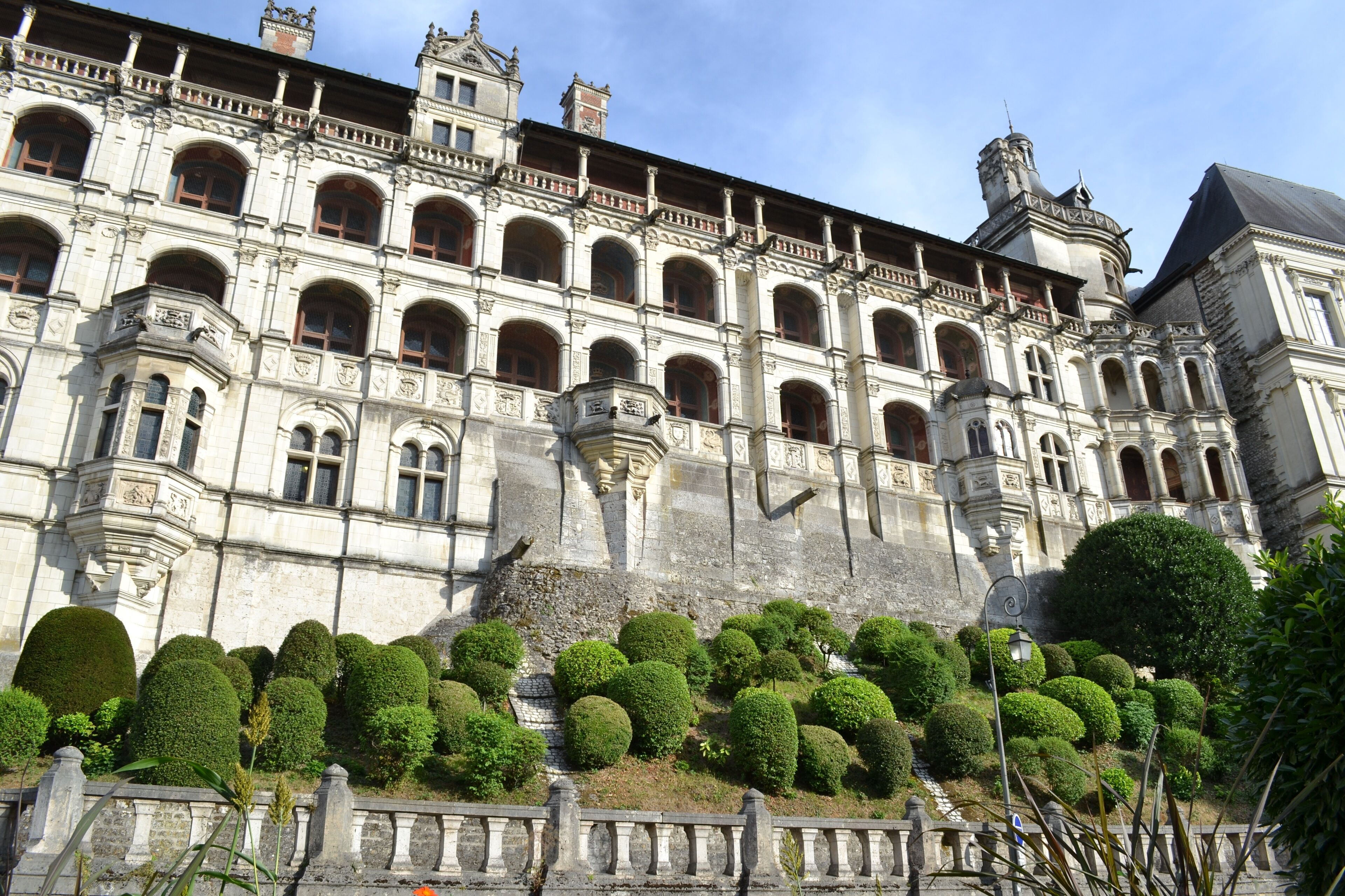 Um edifício histórico com arquitetura ornamentada, várias janelas em arco e um jardim bem cuidado com arbustos bem aparados.