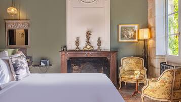 Un dormitorio con una cama grande, una chimenea, dos sillones, una mesita auxiliar, un espejo y un cuadro en la pared.