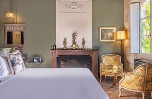Un dormitorio con una cama grande, una chimenea, dos sillones, una mesita auxiliar, un espejo y un cuadro en la pared.