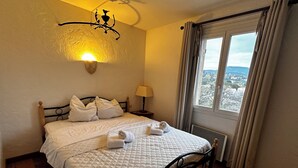 A neatly made bed with white linens and a quilted duvet, a bedside lamp, and a view of the outdoors through a window with curtains. - Family house - Country house - Holiday home (Grimaud)