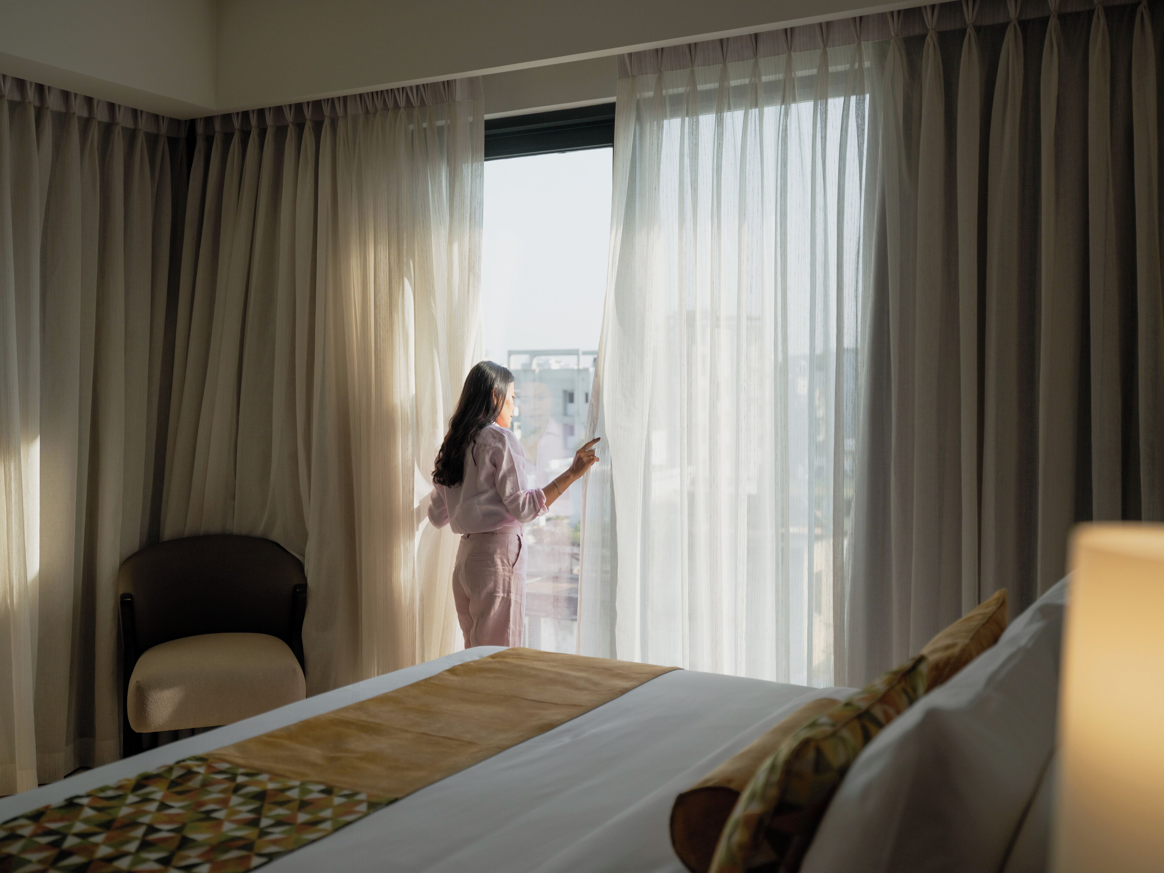 A hotel room with a large bed, a chair, and a view through sheer curtains.