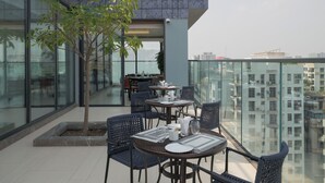A modern outdoor dining area with glass railings, a tree, and a view of the cityscape. - W Residence Living (Dhaka)