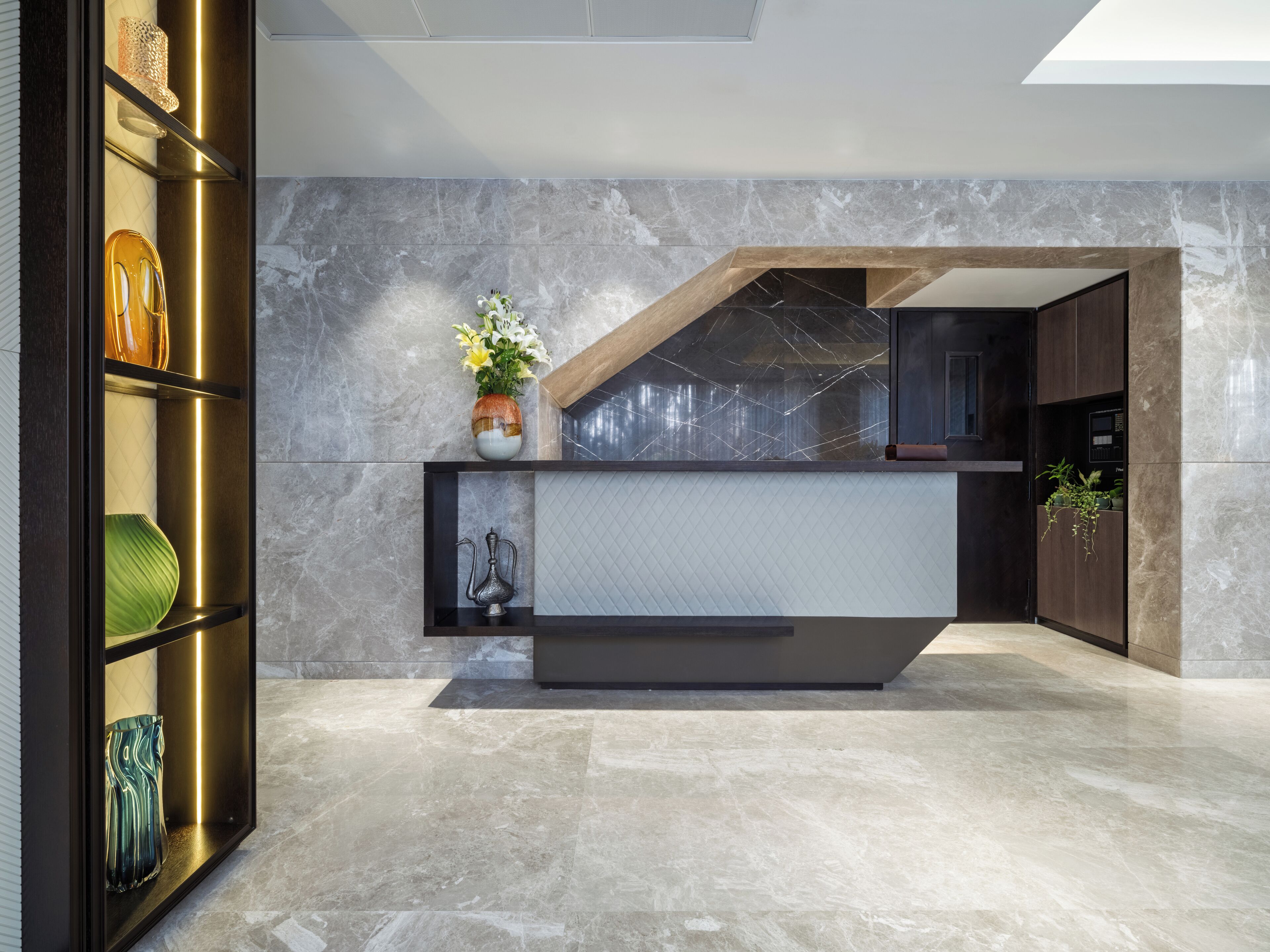 A modern reception desk with a marble wall backdrop, a glass display case with decorative items, and a plant on the counter.