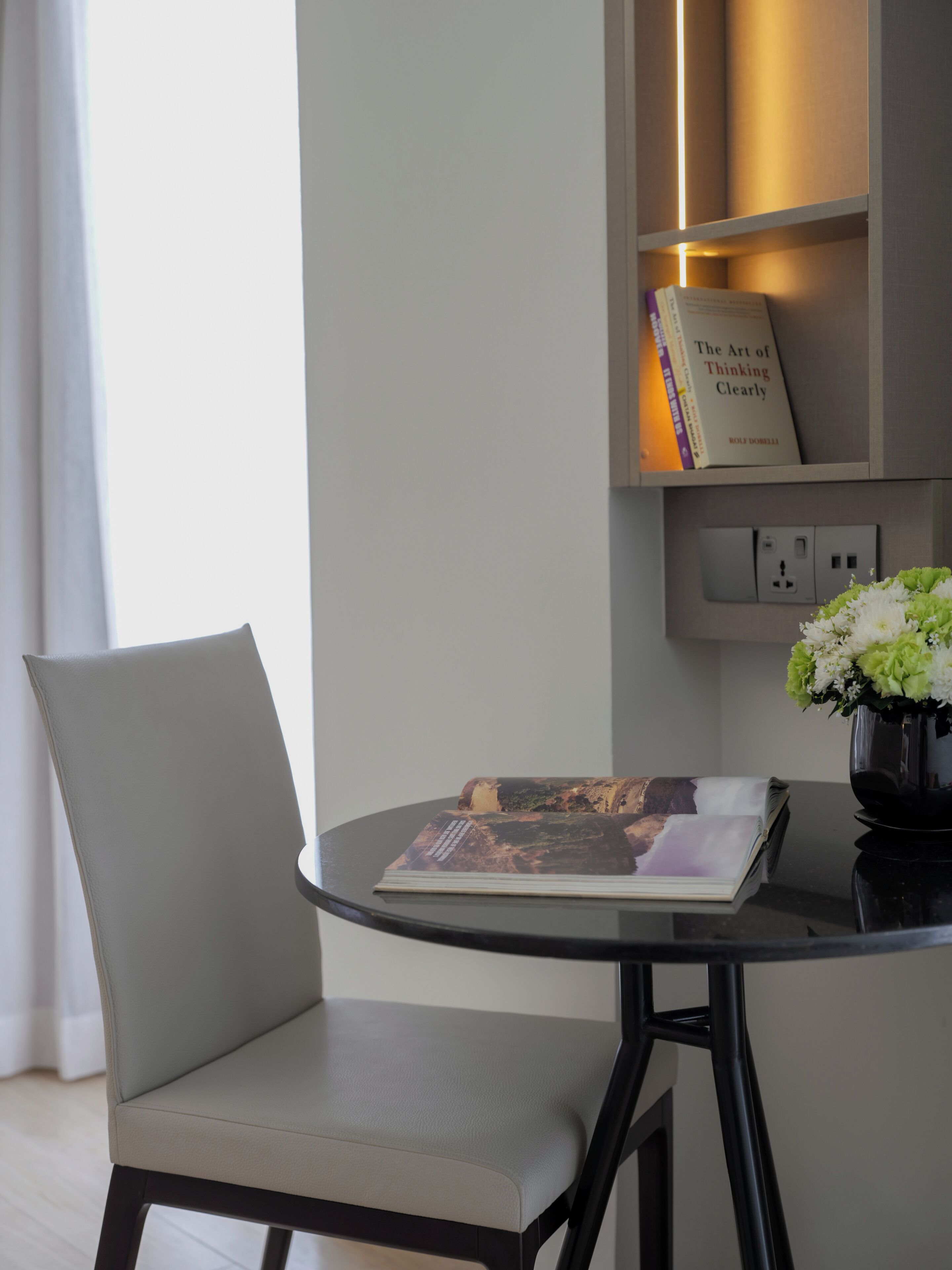 A modern room with a round table, a single chair, a bookshelf with a book titled "The Art of Thinking Clearly," and a vase with flowers.