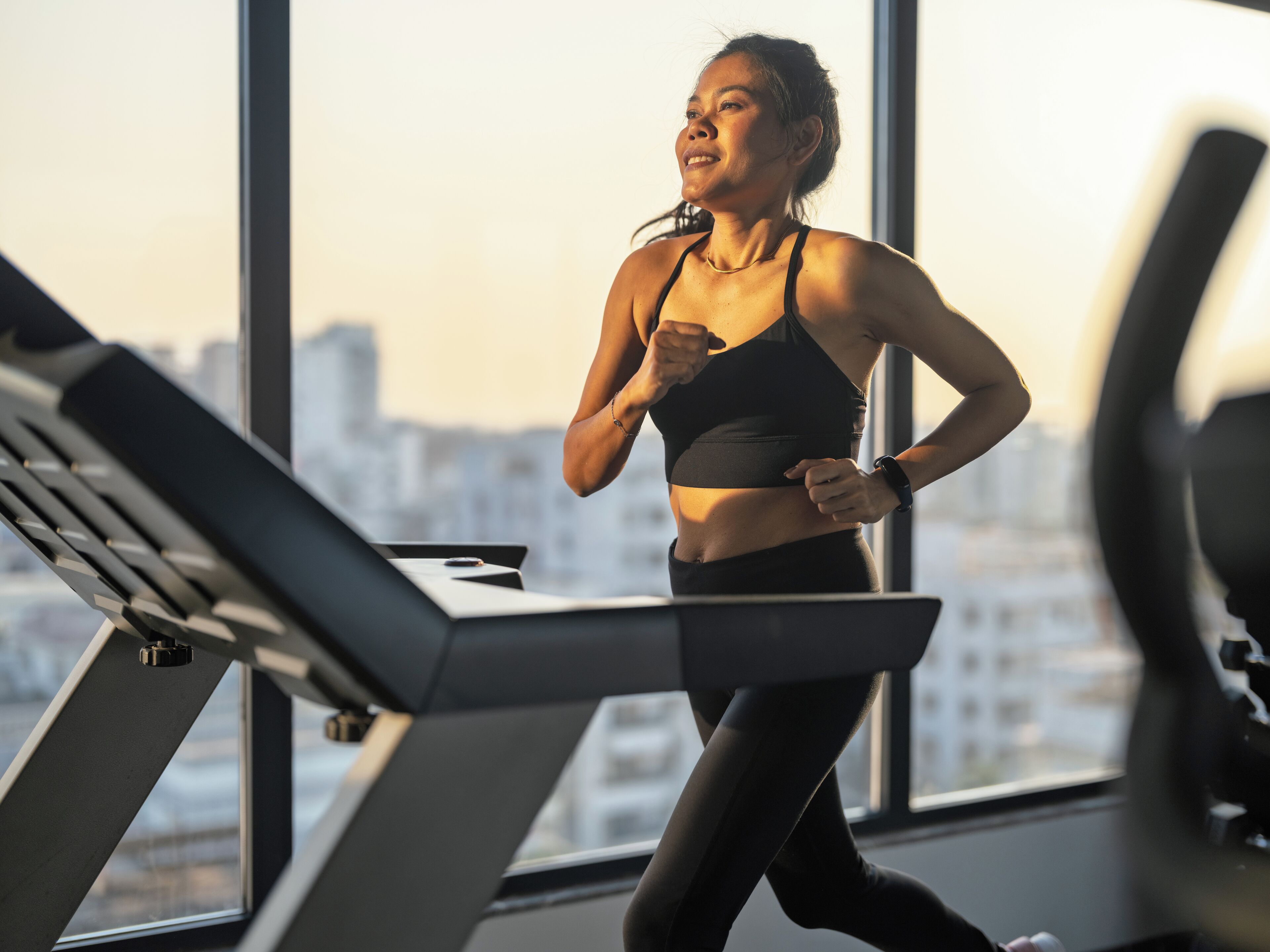 A woman running on a treadmill in a gym with large windows and a city view.