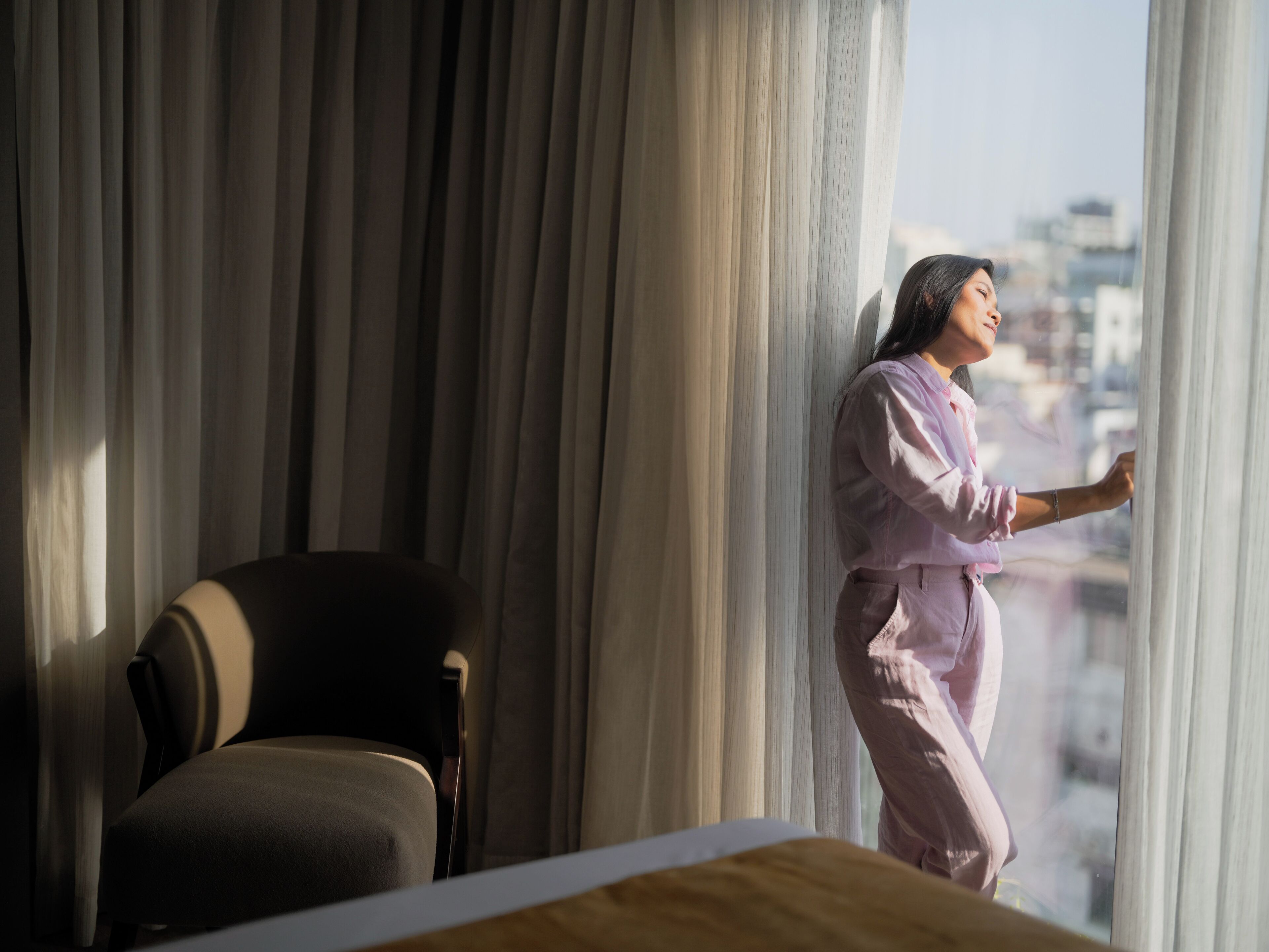 A person standing by a window with curtains, a chair, and a table in a room with a view of buildings outside.