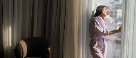 A person standing by a window with curtains, a chair, and a table in a room with a view of buildings outside.