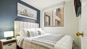 A neatly arranged bedroom with a bed, bedside table, lamp, and a cityscape painting on the wall. - Tourist neighborhood 1 block from the subway (Santiago)