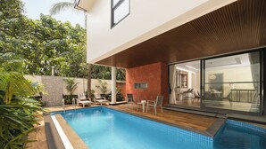 Un área de piscina exterior moderna con terraza de madera, sillas de descanso y puerta de vidrio que da a un espacio interior.