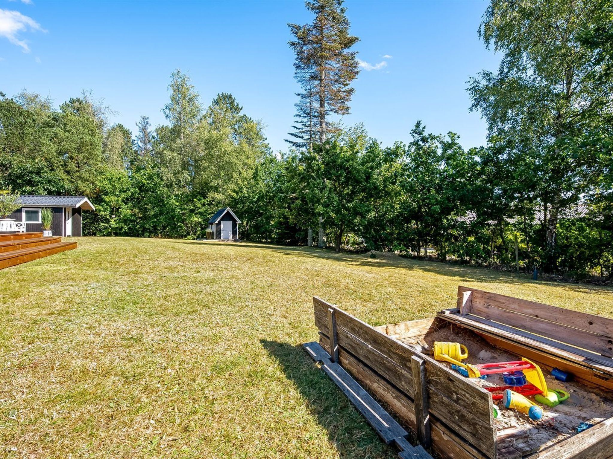 Une cour arrière verdoyante avec une structure de jeu en bois, un petit abri de jardin et un grand arbre.