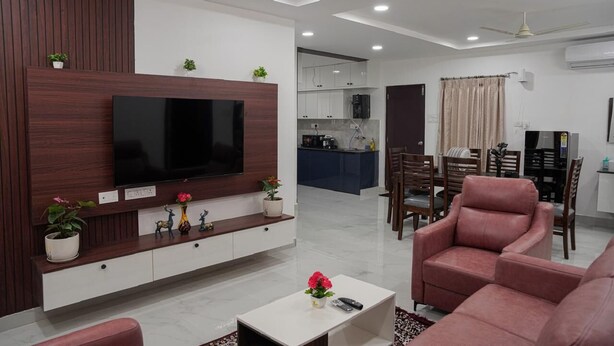 Una sala de estar moderna con una televisión de pantalla plana montada en una pared de madera, un comedor con mesa y sillas, y una cocina al fondo.