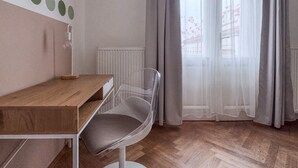 Una habitación con un escritorio de madera, una silla transparente, un radiador y una ventana con cortinas translúcidas.