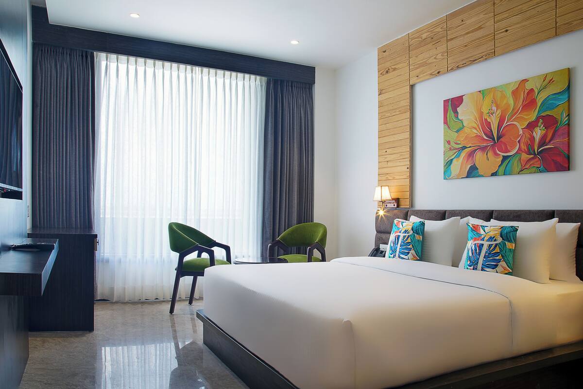 Habitación de hotel con una cama grande, dos sillas verdes, un escritorio y un cuadro colorido en la pared.