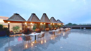 A rooftop terrace with multiple thatched-roof structures, outdoor seating, and a reflective floor. - TreeHouse Velis Sonipat (Ganaur)